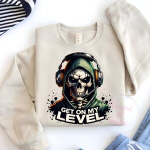 May include: A beige sweatshirt with a graphic print of a skull wearing a green hooded sweatshirt and headphones. The text "Get on my level" is printed below the skull.