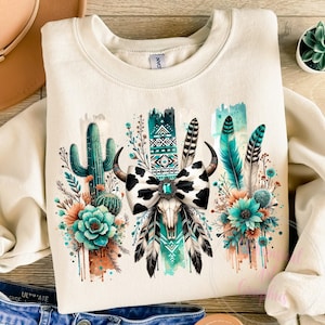 May include: A beige sweatshirt with a graphic design featuring a cow skull with a black and white bow, turquoise feathers, and cactus plants. The design is surrounded by watercolour-style splashes of turquoise, green, and orange.