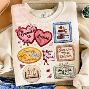 May include: Cream-colored sweatshirt with embroidered patches featuring hearts, books, and phrases like "Enemies to Lovers" and "Just One More Chapter." The design includes a sun, cloud, and open book with candles.