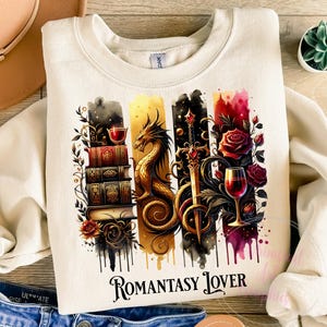 May include: A cream colored sweatshirt with a watercolor design featuring a dragon, a sword, a wine glass, and a stack of books. The design is surrounded by roses and vines. The text "Romantasy Lover" is printed below the design.