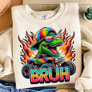 Może przedstawiać: Biała bluza z kolorowym nadrukiem przedstawiającym dinozaura w okularach przeciwsłonecznych i czapce, prowadzącego monster trucka. Pod truckiem znajduje się napis "BRUH".