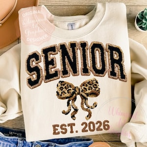以下が含まれることがあります： クリーム色のスウェットシャツ。大きな黒と茶色の文字で「SENIOR」と書かれています。ヒョウ柄のリボンが下にあり、「EST. 2026」がプリントされています。カジュアルなスタイルのスウェットシャツです。