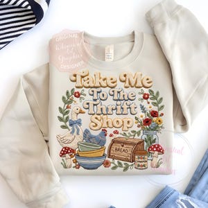 Puede incluir: Sudadera color crema con la frase "Take Me To The Thrift Shop" en una fuente retro, rodeada por una corona floral e ilustraciones de un ganso, una gallina, cuencos y setas. Un dise&ntilde;o de inspiraci&oacute;n vintage.