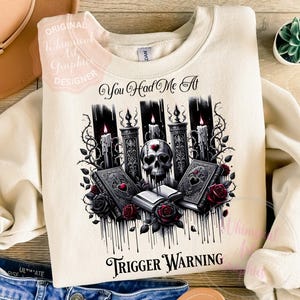 May include: Cream-colored sweatshirt with a graphic design featuring a skull, candles, books, and roses. The text "You Had Me At Trigger Warning" is printed on the front. The design is in black, white, and red.