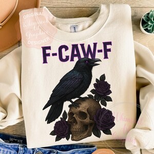 May include: Cream-coloured sweatshirt with a graphic featuring a black raven perched on a skull, surrounded by purple roses. The text "F-CAW-F" is above the raven. A whimsical design for those who love gothic and spooky themes.