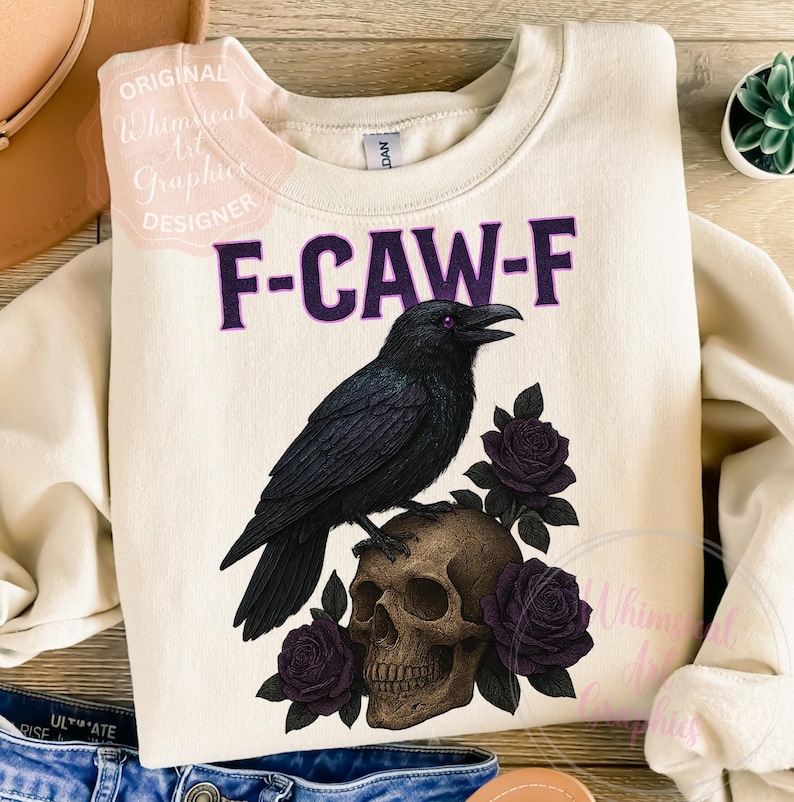 May include: Cream-coloured sweatshirt with a graphic featuring a black raven perched on a skull, surrounded by purple roses. The text "F-CAW-F" is above the raven. A whimsical design for those who love gothic and spooky themes.