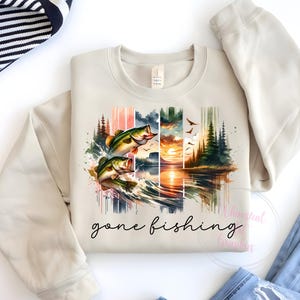 May include: A beige sweatshirt with a watercolor design of two bass jumping out of the water. The design includes a sunset, trees, and the text "gone fishing".