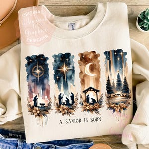 Può includere: Felpa color crema con un design ad acquerello della scena della Natività. Il design include una stella, una luna crescente e sagome di figure, con il testo "A Savior Is Born" in basso.