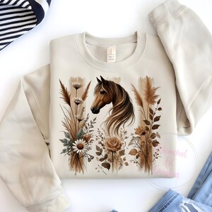May include: A beige sweatshirt with a brown horse and floral design. The horse is facing left and has a brown mane and tail. The flowers are in shades of brown and white.