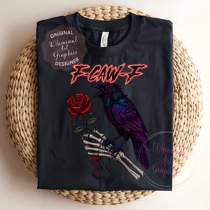 May include: Black t-shirt with a purple raven on a skeletal hand holding a red rose. The text "F-CAW-F" is in red, with "Original Whimsical Art Graphics Designer" and "Whimsical Art Graphics" logos. The shirt is displayed on a woven surface.