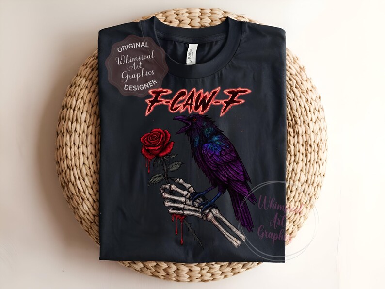May include: Black t-shirt with a purple raven on a skeletal hand holding a red rose. The text "F-CAW-F" is in red, with "Original Whimsical Art Graphics Designer" and "Whimsical Art Graphics" logos. The shirt is displayed on a woven surface.