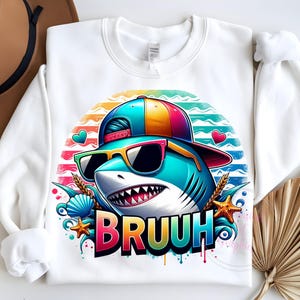 Puede incluir: Sudadera blanca con un tibur&oacute;n de dibujos animados con gafas de sol y una gorra colorida. El tibur&oacute;n tiene una sonrisa con dientes y la palabra "BRUUH" en colores vibrantes. El dise&ntilde;o incluye corazones, conchas y estrellas de mar.