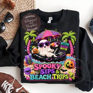 May include: Black sweatshirt with a colorful graphic featuring a ghost wearing sunglasses, a flamingo, and a black cat in a pool. The design includes palm trees, a pumpkin, and the text "Spooky Sips & Beach Trips."