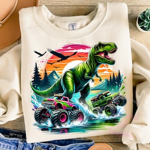 May include: Cream-coloured sweatshirt with a colourful graphic of a green Tyrannosaurus Rex and two monster trucks racing through water. The design includes a sunset, trees, and birds, creating a dynamic scene.