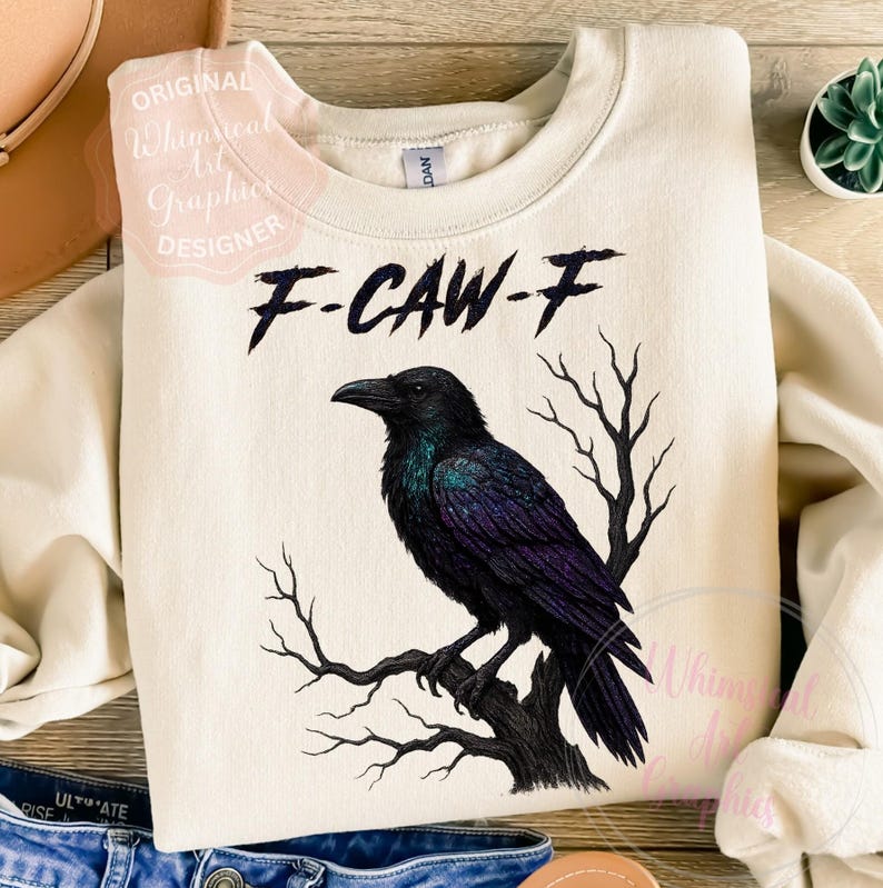 May include: A cream-coloured sweatshirt with a black raven design. The raven, with iridescent purple and green feathers, is perched on a tree branch. The text "F-CAW-F" is printed above the raven. Design by "Original Whimsical Art Graphics Designer".