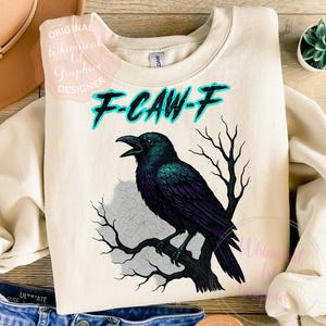 May include: A cream-coloured sweatshirt featuring a black crow graphic perched on a branch. The crow has iridescent green and purple feathers. The text "F-CAW-F" is above the crow in turquoise. The sweatshirt is a casual style.