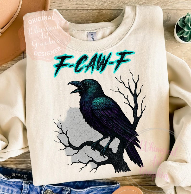 May include: A cream-coloured sweatshirt featuring a black crow graphic perched on a branch. The crow has iridescent green and purple feathers. The text "F-CAW-F" is above the crow in turquoise. The sweatshirt is a casual style.