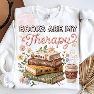 May include: White sweatshirt with the embroidered phrase "BOOKS ARE MY Therapy" and a stack of books with floral accents. The books are labeled "STORIES", "YARN & THREAD", "FLORA & FIBERS", and "QUIET MOMENTS". A coffee cup is also featured.