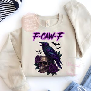 May include: A cream-coloured sweatshirt with a graphic of a purple raven on a skull, with purple roses and bats. The text "F-CAW-F" is in purple. A casual, gothic-themed design.