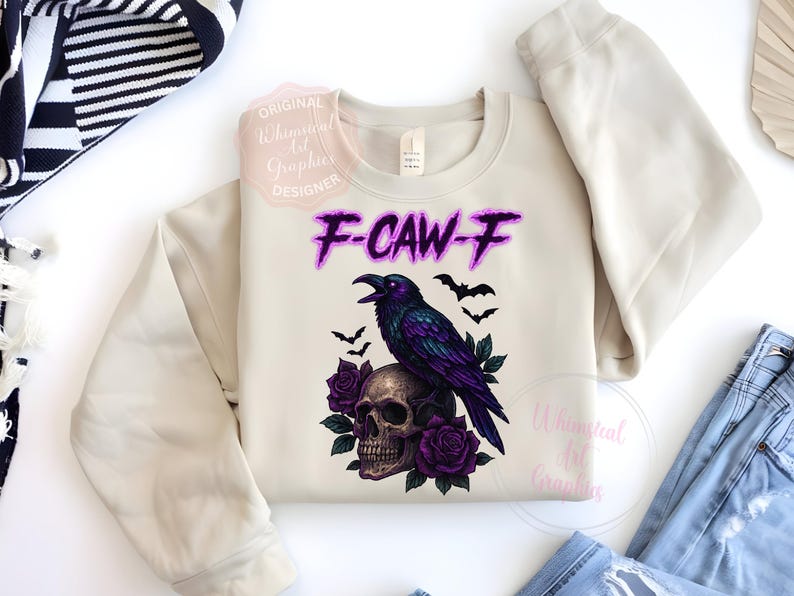 May include: A cream-coloured sweatshirt with a graphic of a purple raven on a skull, with purple roses and bats. The text "F-CAW-F" is in purple. A casual, gothic-themed design.