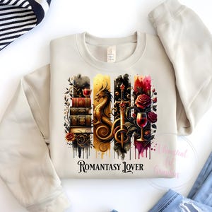 May include: A beige sweatshirt with a graphic print of a dragon, a sword, a glass of wine, and a stack of books. The graphic is surrounded by watercolor-like drips. The text "Romantasy Lover" is printed below the graphic.