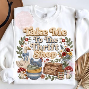Puede incluir: Sudadera blanca con la frase "Take Me To The Thrift Shop" en una fuente retro. El dise&ntilde;o incluye un ganso, una gallina, cuencos, pan, flores y setas, rodeados por una corona floral.