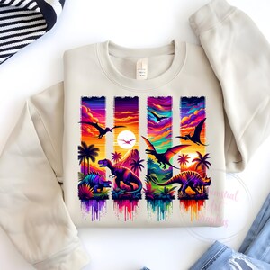 May include: A cream-colored sweatshirt featuring a vibrant graphic of dinosaurs and flying reptiles against a sunset backdrop. The design includes palm trees and a colorful sky, with the text "Essential Life Designs" at the bottom.