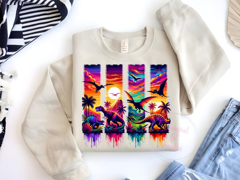 May include: A cream-colored sweatshirt featuring a vibrant graphic of dinosaurs and flying reptiles against a sunset backdrop. The design includes palm trees and a colorful sky, with the text "Essential Life Designs" at the bottom.