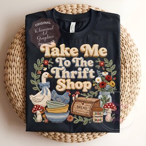 Puede incluir: Camiseta negra con la frase "Take Me To The Thrift Shop" en una fuente retro, rodeada de ilustraciones de un ganso, una gallina, setas, flores y cuencos. La camiseta tiene una est&eacute;tica caprichosa y vintage.