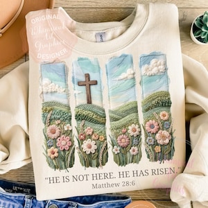 May include: Cream-coloured sweatshirt with an embroidered design. It features a wooden cross, rolling hills, blue skies, and colourful flowers. The text "HE IS NOT HERE. HE HAS RISEN." Matthew 28:6 is printed below. A comfortable, casual style.