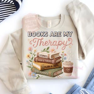 May include: Beige sweatshirt with the embroidered text "BOOKS ARE MY Therapy" and a stack of books with floral accents. The books are labeled "STORIES", "YARN & THREAD", "FLORA & FIBERS", and "QUIET MOMENTS". A coffee cup is also featured.