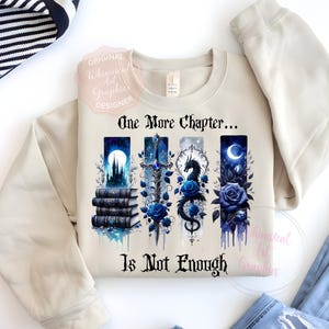 May include: Beige sweatshirt with a graphic design featuring the text "One More Chapter... Is Not Enough." The design includes illustrations of books, a castle, a sword, a dragon, and blue roses. The sweatshirt is paired with blue jeans.