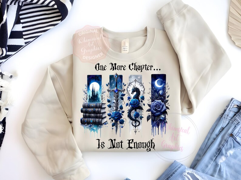 May include: Beige sweatshirt with a graphic design featuring the text "One More Chapter... Is Not Enough." The design includes illustrations of books, a castle, a sword, a dragon, and blue roses. The sweatshirt is paired with blue jeans.