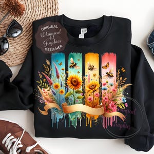 May include: Black sweatshirt featuring a colorful floral and bee design. The design includes sunflowers, wildflowers, and butterflies on vertical watercolor-style panels. A gold ribbon banner is across the bottom. The text "Original Whimsical Art Graphics Designer" is on the design.