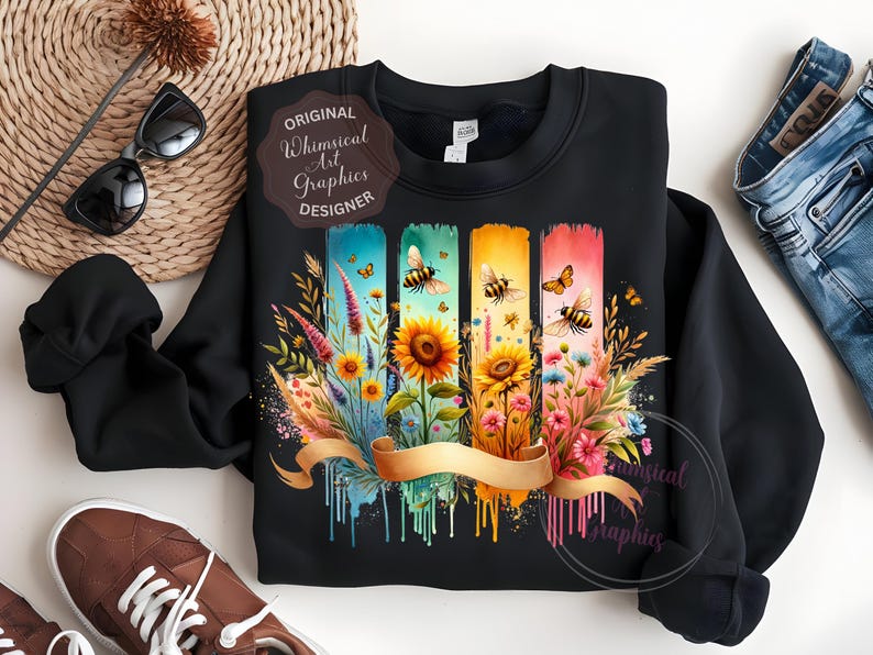 May include: Black sweatshirt featuring a colorful floral and bee design. The design includes sunflowers, wildflowers, and butterflies on vertical watercolor-style panels. A gold ribbon banner is across the bottom. The text "Original Whimsical Art Graphics Designer" is on the design.