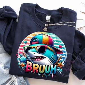 Puede incluir: Sudadera azul marino con un tibur&oacute;n de dibujos animados con gafas de sol y una gorra colorida. El gr&aacute;fico del tibur&oacute;n incluye la palabra "BRUUH" en colores arco&iacute;ris. El dise&ntilde;o incorpora corazones, conchas marinas y estrellas de mar. Un collar plateado con un colgante est&aacute; sobre la sudadera.