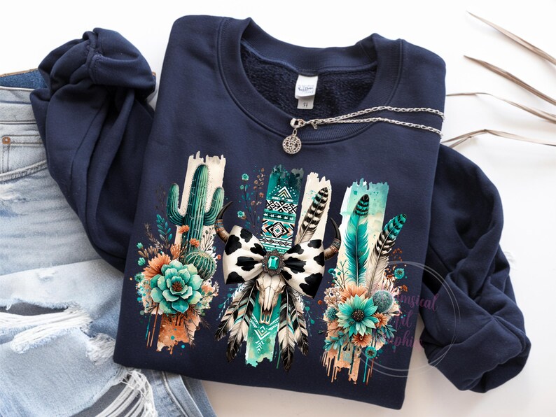 May include: Navy blue sweatshirt featuring a graphic print of a cow skull with a turquoise and white bow, surrounded by cactus, feathers, and flowers.