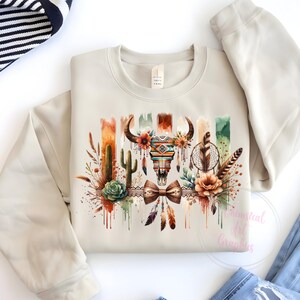 May include: A beige sweatshirt with a watercolor design featuring a skull with feathers, dreamcatcher, and cacti. The design is in shades of brown, orange, green, and blue.