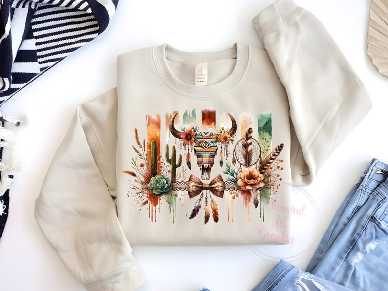 May include: A beige sweatshirt with a watercolor design featuring a skull with feathers, dreamcatcher, and cacti. The design is in shades of brown, orange, green, and blue.