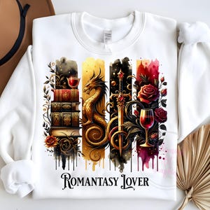 May include: White sweatshirt with a graphic design of a dragon, a sword, a glass of wine, and a stack of books. The graphic is surrounded by watercolor-like splatters of red, yellow, and blue. The text "Romantasy Lover" is printed below the graphic.