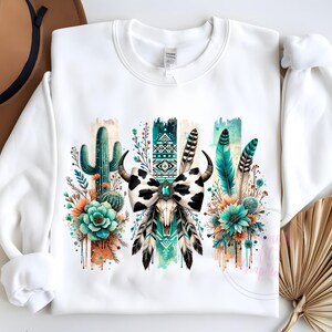 May include: White sweatshirt featuring a graphic print of a cow skull with a bow, cactus, and feathers in shades of green, blue, and orange.
