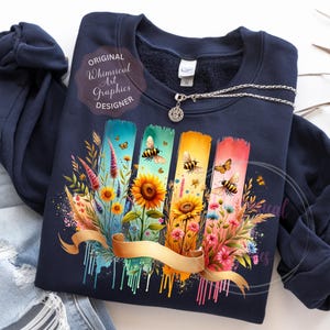 May include: Navy blue sweatshirt featuring a colorful floral design with bees, sunflowers, and butterflies. The design includes vertical brushstrokes in blue, green, yellow, and pink, with a gold ribbon. The text "Original Whimsical Art Graphics Designer" is also visible.