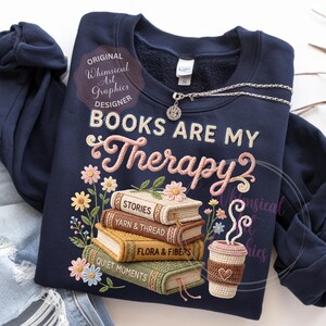 May include: Navy blue sweatshirt with the embroidered phrase "BOOKS ARE MY Therapy" and a stack of books with floral accents. A crocheted coffee cup and a silver necklace are also visible.