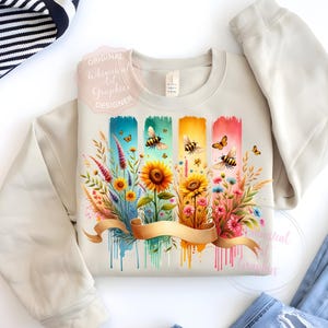 May include: Beige sweatshirt with a colorful floral and bee design. The graphic features sunflowers, wildflowers, and bees on a watercolor background with a gold ribbon. The text "Original Whimsical Art Graphics Designer" is visible.