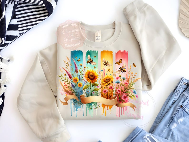 May include: Beige sweatshirt with a colorful floral and bee design. The graphic features sunflowers, wildflowers, and bees on a watercolor background with a gold ribbon. The text "Original Whimsical Art Graphics Designer" is visible.