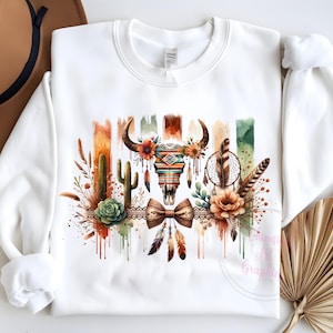 May include: White sweatshirt with a watercolor design of a skull with feathers, cacti, and flowers. The skull is decorated with geometric patterns and a bow tie.