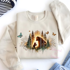 May include: A beige sweatshirt with a graphic print of a mother bear and her cub standing in a forest with butterflies. The graphic is in shades of brown, green, and blue.