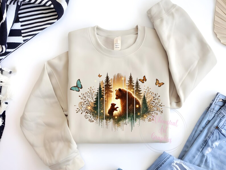 May include: A beige sweatshirt with a graphic print of a mother bear and her cub standing in a forest with butterflies. The graphic is in shades of brown, green, and blue.