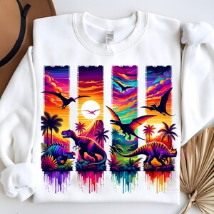 May include: White sweatshirt featuring a colorful dinosaur design. The graphic includes various dinosaurs, palm trees, and flying pterodactyls set against a vibrant sunset backdrop. The design is rendered in shades of purple, orange, and blue.