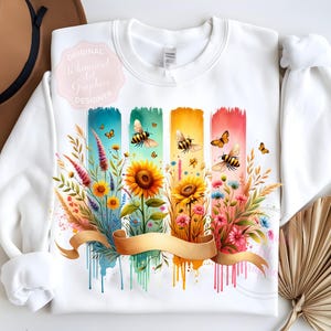 May include: White sweatshirt featuring a colorful floral and bee design. The design includes sunflowers, wildflowers, and butterflies, with a gold ribbon across the bottom. The words "Original Whimsical Art Graphics Designer" are on the upper left.